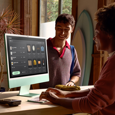 An employee sitting behind the counter at a shop that sells surfboards and other items uses an iMac to complete a purchase for a customer on the other side of the counter.